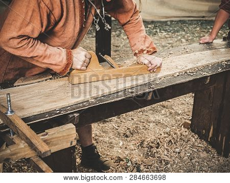Carpenter In Medieval Cotton Clothes Working With A Wood By Plane. Man Manually Hews A Wooden Board 
