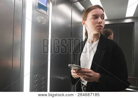 Businesswoman Is Taking A Look In The Elevator Using Her Smartphone And Headphones. Portrait Of A Fe