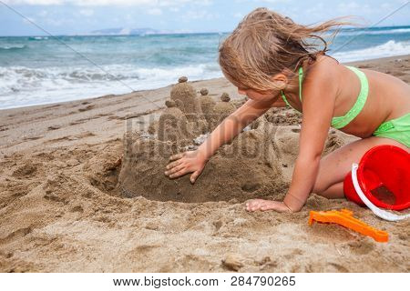 Little girl wearing green swimsuitbuilding a sand cactle on a sandy beach in Crete, Greece. Family summer vacation concept