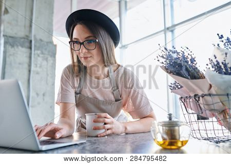 Young woman in workwear holding mug with herbal tea while bending over table in front of l aptop