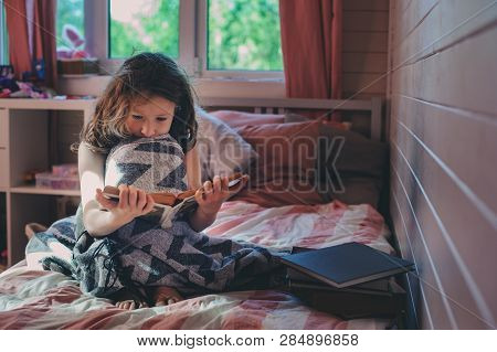 Thoughtful Kid Girl Reading Book Alone In Her Room In The Veening Or Early Morning