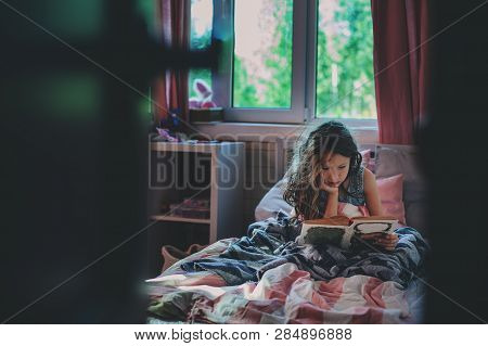 Thoughtful Kid Girl Reading Book Alone In Her Room In The Veening Or Early Morning
