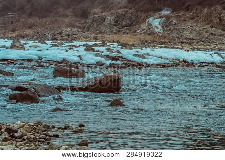 Rapidly Flowing River With Rocky Riverbed And Rocky Shoreline Covered With Sheets Of Ice Snow.