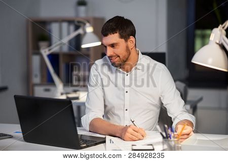 business, deadline and people concept - businessman with laptop computer and papers working at night office
