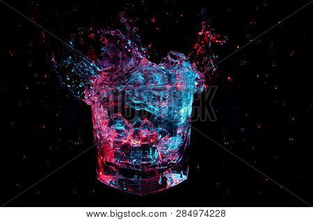Huge Splash Of Transparent Liquid Glowing In Blue And Red Lights In A Rocks Glass Isolated On A Blac