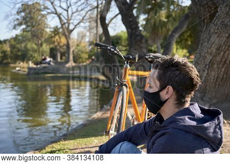 Young Man With A Cloth Mask Resting With His Bike By A Lake In A Park In Buenos Aires, Argentina, On