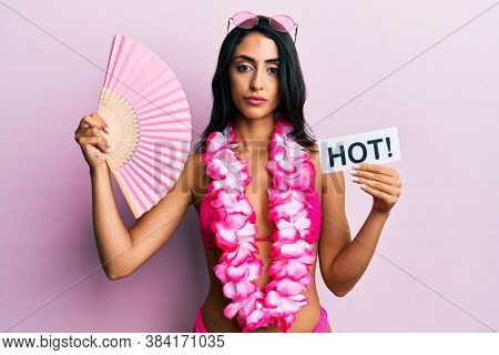 Beautiful hispanic woman wearing bikini and handfan holding hot word relaxed with serious expression on face. simple and natural looking at the camera. 