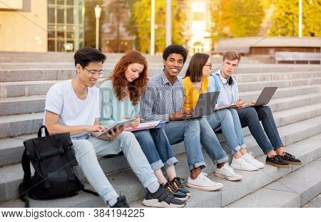 Multiethnic Students Learning Together Using Gadgets Sitting On Steps Outdoors At University Entranc