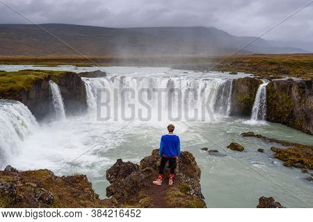 Young Hiker Standing At The Godafoss Waterfall In Iceland. Godafoss Means The Waterfall Of The Gods 