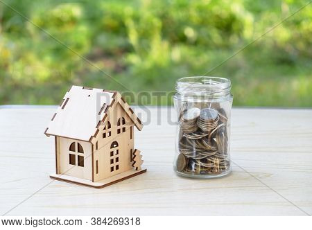 A Small Wooden House And A Glass Jar With Coins. The Concept Of Buying, Selling, Renting Real Estate