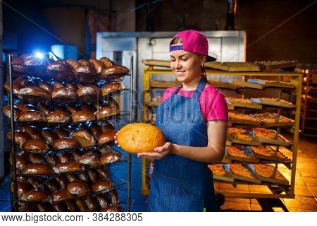 A Baker Girl Takes Hot Bread In A Bakery Against The Background Of Shelving With Bread. Industrial P