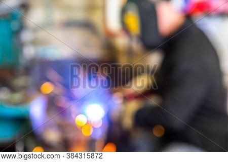 Blurred Background Of Electric Welding Process. Worker In Welding Mask. Yellow Sparks Fly To The Sid