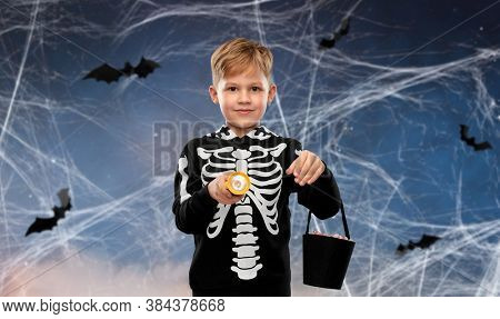halloween, holiday and childhood concept - little boy in black costume of skeleton with bucket of candies and flashlight trick-or-treating over night sky with bats and cobweb on background