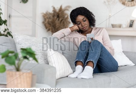 Apathy Concept. Depressed African Woman Sitting With Smartphone On Sofa At Home, Looking At Device S