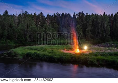 Traditional Midsummer Ligo Celebration. Huge Bonfire On The River Shore. Scenic Landscape Of Forest 