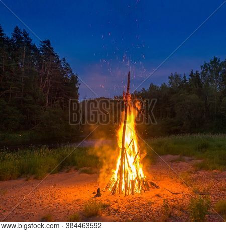 Midsummer Ligo Celebration. Huge Fire On The River Shore. Scenic Landscape Of Forest At Twilight.