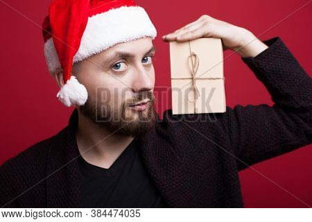 Close Up Photo Of A Bearded Caucasian Man With Santa Hat Holding A Gift On His Shoulders Posing On A
