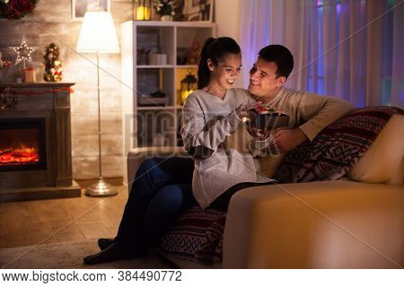 Young Couple On Christmas Day Sitting On Couch In Dark Living Room With Fireplace In The Background.