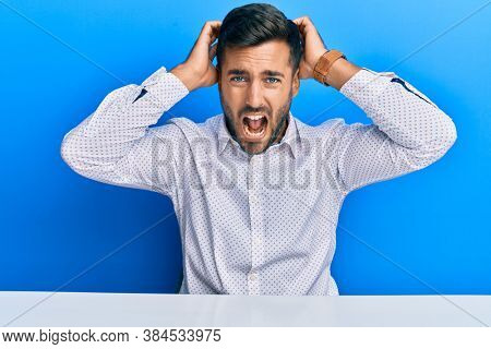 Handsome hispanic man wearing business clothes sitting on the table crazy and scared with hands on head, afraid and surprised of shock with open mouth 