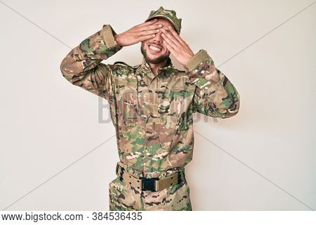 Young caucasian man wearing camouflage army uniform covering eyes with hands smiling cheerful and funny. blind concept. 