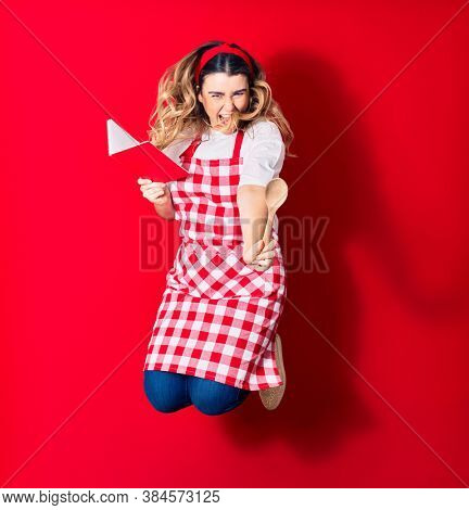 Young beautiful blonde cook woman wearing apron smiling happy. Jumping with smile on face holding recipe book and spoon over isolated red background
