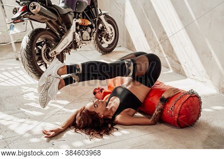 Woman With Multiple Hobbies Posing Near Her Motorcycle And Posing Leaning On Punching Bag In Trainin