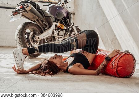 Woman With Multiple Hobbies Posing Near Her Motorcycle And Posing Leaning On Punching Bag In Trainin
