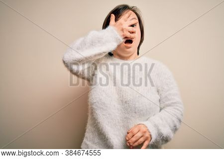 Young down syndrome woman standing over isolated background peeking in shock covering face and eyes with hand, looking through fingers with embarrassed expression.