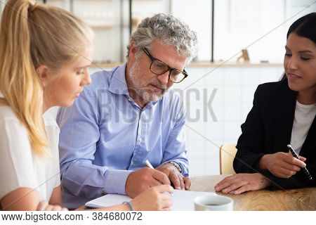 Focused Young And Mature Customers Meeting With Agent And Signing Insurance Agreement. Group Of Busi