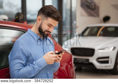 Young Handsome Man Looking Away Thoughfully While Using Smart Phone At Car Dealership, Copy Space