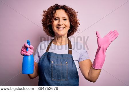 Middle age curly hair woman cleaning doing housework wearing apron and gloves using spayer very happy and excited, winner expression celebrating victory screaming with big smile and raised hands