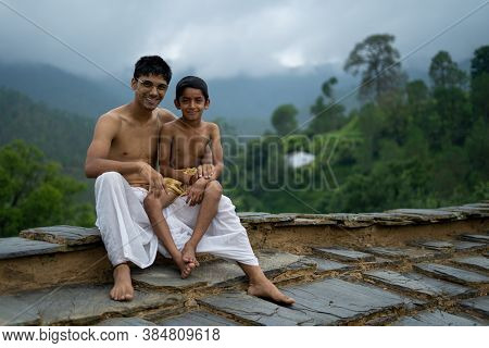 A Young Man Sitting On The Roof With His Younger Brother,both Wearing Dhoti, Smiling Into The Camera