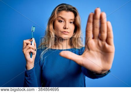 Young beautiful blonde woman using teeth brush over isolated blue background with open hand doing stop sign with serious and confident expression, defense gesture