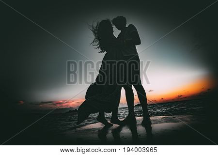 Silhouettes of young couple: man and woman standing on wet ocean pier and hugging each other looking into eyes during dramatic blue and orange windy sunset on Maldives resort with water in background