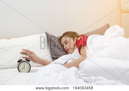 asian young woman in red pajamas sleeping on bed and turning off alarm clock times at 7 o'clock in bedroom at the morning selective focussunlight effect