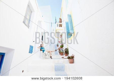 Young attractive woman wearing shorts and sandals, sitting on chair between white traditional greek houses and enjoying colours of authentic greek village full of flowers - Greece holidays concept