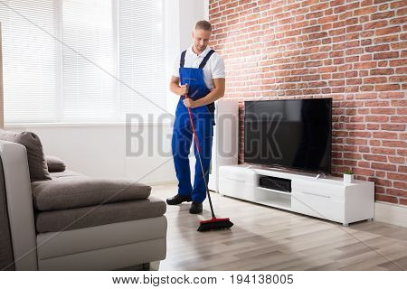 Smiling Male Janitor In Uniform Sweeping Floor With Broom At Home