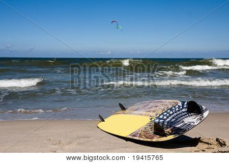 Junta de Kiteboarding tendido en una playa. Dos kiteboardes en un segundo plano.