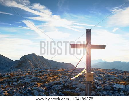 Modest Wooden Cross Raised On Rocky Alpine Mountain Summit . Sharp Rocky Peak. Gentle Clouds  In Blu