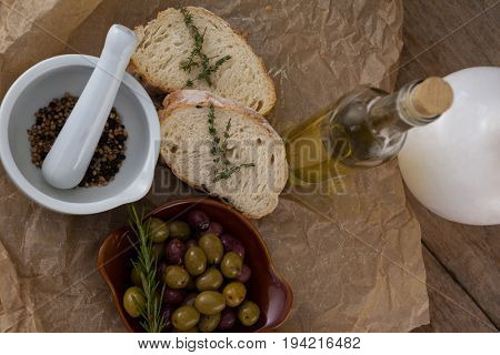 Directly above shot of olives with oil bottle and bread by spices in mortar pestle on crumbled paper at table