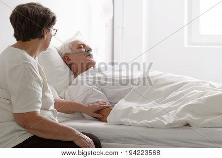 Senior man in a coma laying on hospital bed and his wife sitting next to him