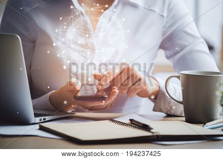 Woman using smartphone with glowing spehere at workplace with notepad coffee cup laptop and other items on desktop. Networking concept