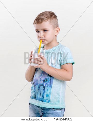 Charming little boy in a blue t-shirt drinks milk from a yellow straw. A cute little boy is holding a glass of milk. Adorable little boy is drinking fresh milk on gray background.