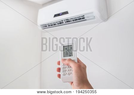 Hand holding remote control, adjusting temperature of air conditioner mounted on a white wall. Indooor comfort temperature