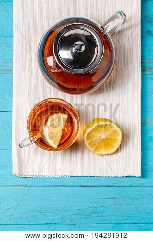 Glass cup of tea with lemon and glass teapot on blue wooden table.