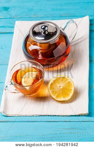 Glass cup of tea with lemon and glass teapot on blue wooden table.