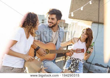 Three young friends having fun at rooftop party playing guitar and singing. Focus on the man playing the guitar