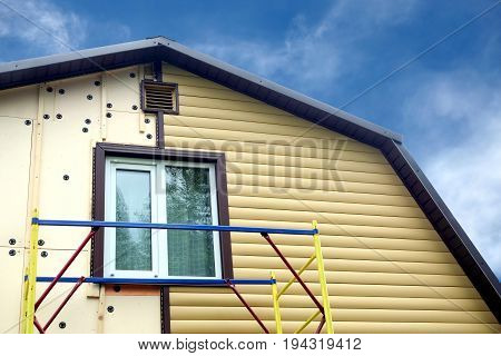 Beige siding panels mounting with scaffolding on top of rural house in sunny day over blue sky front view