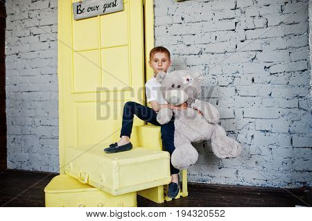Portrait Of A Little Cute Boy Officially Dressed Posing With A Teddy Bear On Yellow Boxes In Grey Ro