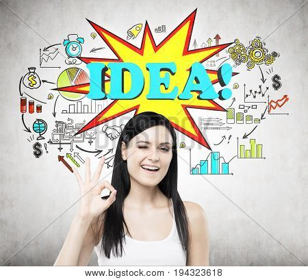 Cheerful black haired woman making an OK sign. Concete wall with a colorful business scheme sketch and a word idea on it. Toned image